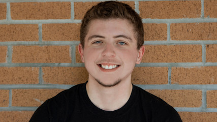 Obraz premium A young pretty pale skinned man with a black shirt, short curly reddish brown hair, blue eyes, a wide soul patch, and a large radiant grin smiles looking into the camera with a slightly lazy right eye