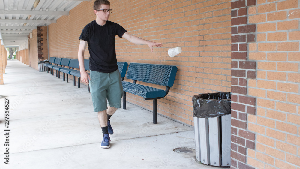 Wall mural A pale skinned young adult man wearing a black shirt and glasses with shorts in a brick hall surrounded by blue benches tosses his empty cup into the trash mod air as he opposes the idea of littering