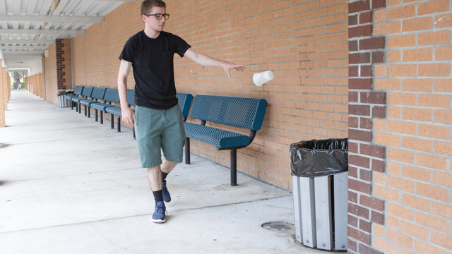 A pale skinned young adult man wearing a black shirt and glasses with shorts in a brick hall surrounded by blue benches tosses his empty cup into the trash mod air as he opposes the idea of littering