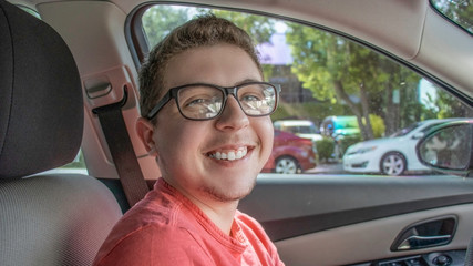 A very young man with blue eyes and short hair smiles big and proudly in his cars driver seat wearing a red shirt and thick rimmed black glasses looking into the camera ready to take on the day ahead.