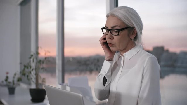 Waist-up shot of grey-haired Caucasian business lady, wearing dark-rimmed glasses, sitting on windowsill in office, with sunset in background, and talking on mobile phone, glancing at laptop screen