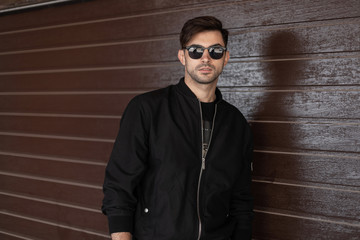 Young attractive hipster man in stylish black jacket in trendy sunglasses with fashionable...
