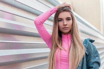 Portrait of a pretty attractive young woman with gorgeous long blond hair in a denim jacket in a pink shirt outdoors near a metallic silver wall. Cute girl. New collection of summer women's clothing.