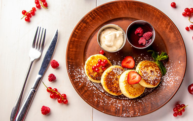 Cottage cheese pancakes, homemade traditional Ukrainian dish ( syrniki ) with berry sauce and sour cream on a brown plate on a white background, horizontal photo