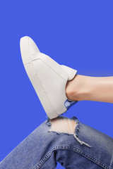 Trendy white eco leather sheakers on bent leg knee in ragged jeans over pastel purple blue background. Colorful main trend backdrop for your magazine, online store and media. Copy space