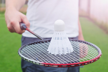 Badminton racket in the hand of a man in a white T-shirt. On the racket lies a shuttlecock. In the background - an open-air stadium.Concept of summer hobby, outdoor sports, entertainment. Copy space