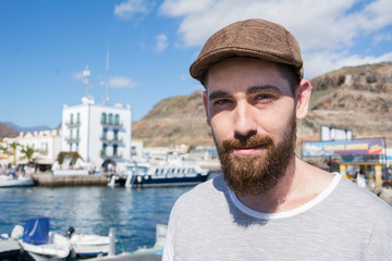 Portrait of a young guy with a beard and hat under the sun in the harbor.