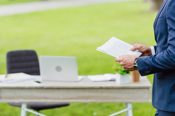 cropped view of young businessman holding newspaper and standing in park near table
