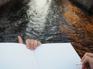 Mockup of person's hand holding blank white notebook preparing to write down his or hers ideas. Close up of female hand holding a pen and opened sketchbook pages on the city canal background.