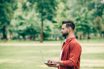 Obraz premium Side view of young man standing in park with laptop in hands and looking away