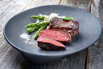 Barbecue dry aged wagyu sirloin beef steak sliced green asparagus and parmesan cheese as closeup on a gray cast iron design plate