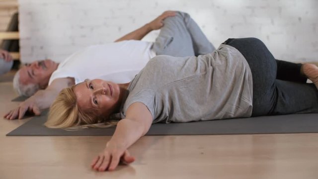 Tracking shot of smiling middle-aged Caucasian husband and wife stretching on mats in fitness studio, and 20-something dark-haired personal trainer sitting with crossed legs and giving instructions
