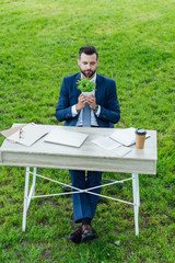 high angle view of young businessman holding plant in flowerpot and sitting behind table with laptop, notebooks and coffee to go