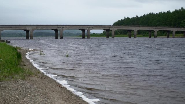 A road bridge over the Blessington Reservoir near Dublin, Ireland. Small waves rippling along a stoney beach. Camera pans right.