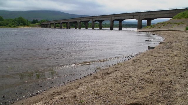 A road bridge over the Blessington Reservoir near Dublin, Ireland. Small waves rippling along a stoney beach. Camera pans left. Wicklow mountains in the background.