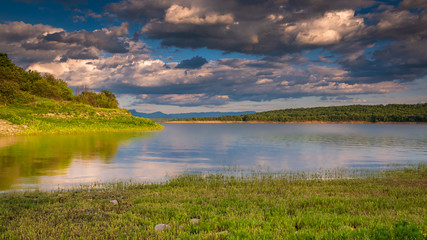 A beautiful scenic of the calm waters of a lake at sunset, with green shores, an island with bush vegetation, distant horizon with woods and mountain range, blue sky with some high contrast clouds