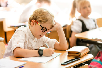 Blonde boy with big black glasses sitting in classroom, studing, smiling. Education on elementary school, first day at school