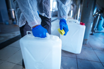 Unrecognizable person technologist in white protective suit handling acid or detergent in chemical...