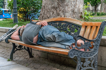homeless man with a big belly sleeping on a wooden bench on a sunny day, next to crutches