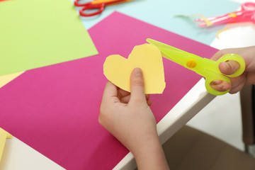 Child cutting out paper heart with plastic scissors at table, closeup. Space for text
