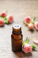 Bottle of rose essential oil and fresh flowers on wooden table, space for text