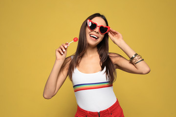 Portrait of beautiful young woman with heart shaped sunglasses and lollipop on color background