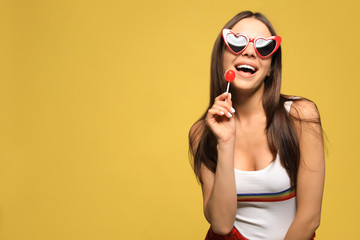 Portrait of beautiful young woman with heart shaped sunglasses and lollipop on color background....