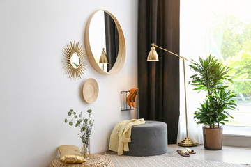 Hallway interior with big round mirror and ottoman chair near white wall
