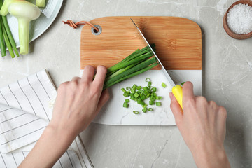 Woman cutting fresh green onion on wooden board at marble table, top view