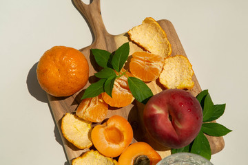 Still Life of Summer Fruit on Wooden Serving Board in Direct Sunlight Next to Glass of Water, Peach, Orange, Nectarine, and Glass of Water, Tan Background, closeup