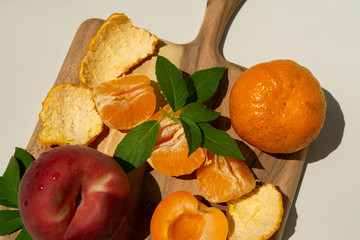 Still Life of Summer Fruit on Wooden Serving Board in Direct Sunlight Next to Glass of Water, Peach, Orange, Nectarine, and Glass of Water, Tan Background, closeup
