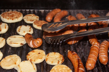 Barbecue food. Hot grill with pork sausages and arepa about to be picked up with a bbq tool. Party with friends concept.