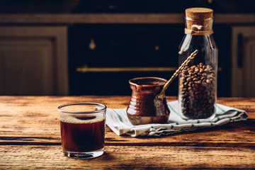 Freshly brewed turkish coffee in drinking glass. Cezve and jar of roasted coffee beans.