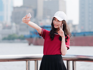 Fashion pretty young girl with black long hair, wearing red T-shirt and white baseball cap selfie with scissors hand pose outdoor, minimalist urban clothing style.