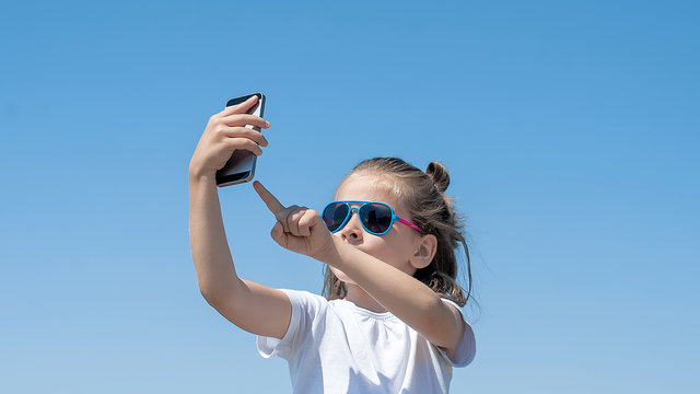Portrait of beautiful happy little girl taking a selfie on the camera phone with blue sky on background. Young smiling child having fun and laughing against clear sky. Summertime and leisure concept