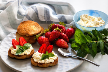 Still life. Sandwiches of bread with white curd, red strawberries, green leaves of mint. On a plate are croutons, ripe berries, a spoon. Cottage cheese in a blue cup. sprigs of mint, linen striped fab