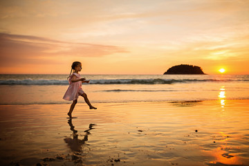 Fototapeta premium Cute little girl walking on the beach. Sunset time. Kid having fun in holiday vacation with back sun light - Youth, lifestyle, travel and happiness concept