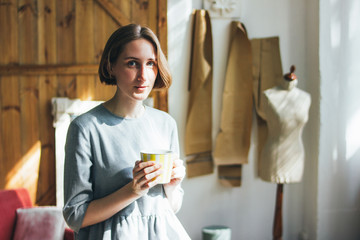 Young woman in simple grey dress with yellow cup sitting on chair in art work studio, full length portrait
