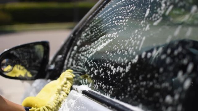 Close-Up Slow motion shot of someone washing a silver family car on a hot summer day with a yellow sponge.