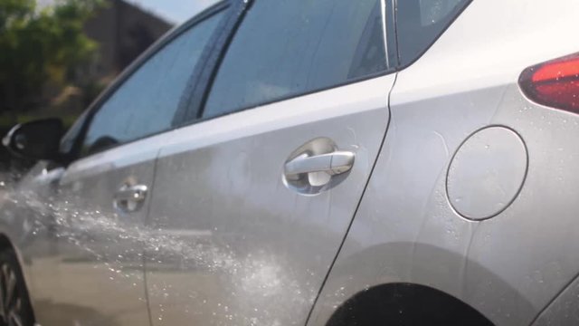 Slow motion shot of someone hosing down a silver family car on a hot summer day.
