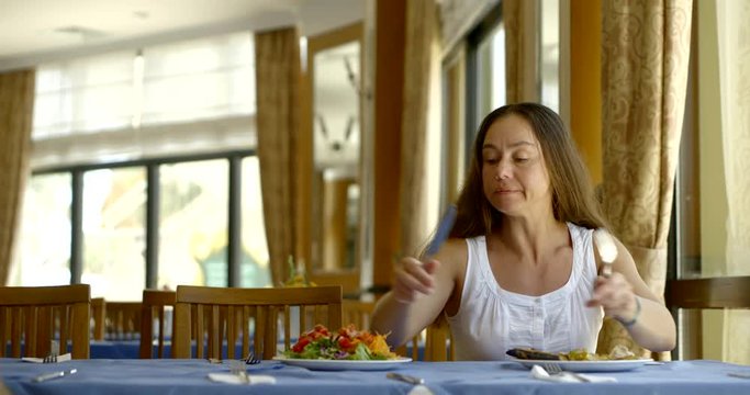 Portrait of a dark-haired middle-aged woman sitting at a table, eating, in front of her hot plate and a plate of salad, in her hands she holds Cutlery - spoon and fork.