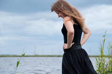 young blonde woman wearing blck dress standing with her hands on her hips near lake, overcast sky