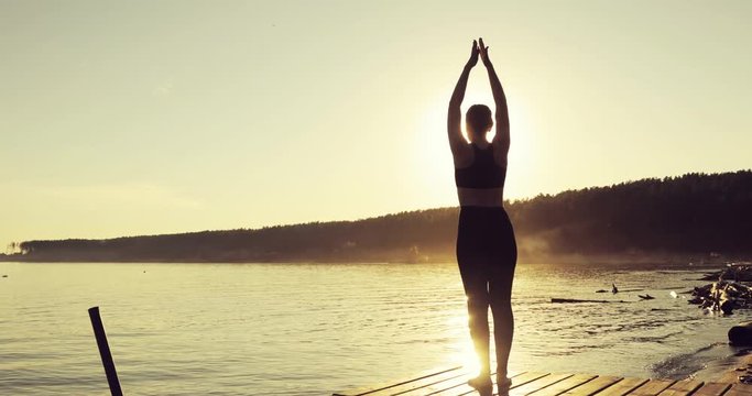 Young woman in sportswear is practicing yoga in tree pose standing on the pier in the yellow rays of the sunset. She is training on nature, back view. Girl is keeping balance on one leg.