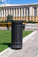 metal round black street bin stands on the sidewalk near the green lawn. Round trash can on the sidewalk.