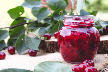 Cherry jam in a glass jar on a background of green leaves on a table of wooden boards.