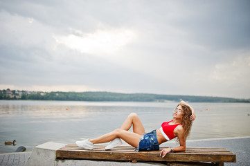 Fototapeta na wymiar Sexy curly model girl in red top, jeans denim shorts, eyeglasses and sneakers posed on bench against lake.