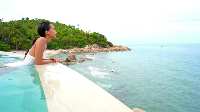 Peaceful scene on tropical island, Asian woman relaxing in infinity pool looking into distance