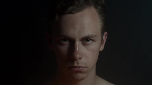 Portrait of an angry disturbing young guy looking into camera in a dark room.