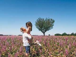 A blond man in a white cotton shirt embraces a brunette girl in a pink dress in a lavender field in Provence in France, Valensol, a heart-shaped tree
