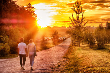 Naklejka na ściany i meble sunset at city park with couple walking along path towards setting sun and town skyline with dramatic sky background landscape street view of people enjoying summer evening authentic lifestyle scene